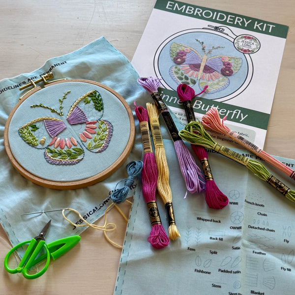 Embroidery kit laid out on a light wood surface, including a butterfly embroidery hoop with finished stitching, multiple colored embroidery floss skeins, green-handled scissors, pattern paper, and an embroidery kit booklet showing a butterfly design.
