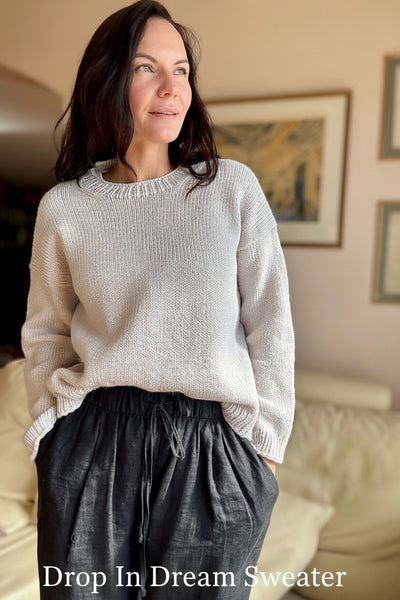A woman wearing a light beige knit sweater and dark gray drawstring pants, standing in a cozy indoor setting with framed art on the wall.