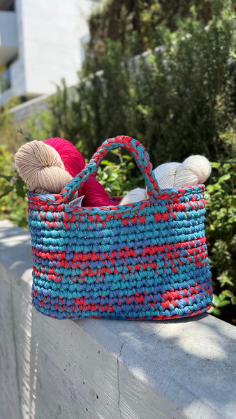A picture of a crochet bag in a blues and red colors with skeins of yarn inside.  The background is of a wall with some greenery behind it. 
