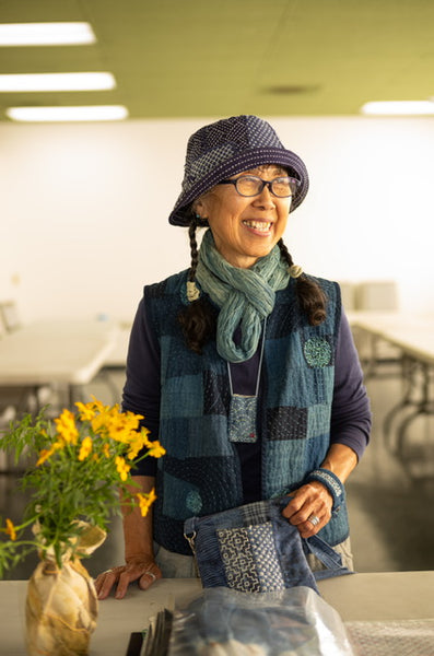 A smiling woman wearing a blue patchwork hat, glasses, a blue scarf, and a blue patchwork vest stands behind a table with a matching blue patchwork pouch and a vase of yellow flowers in a bright indoor room.