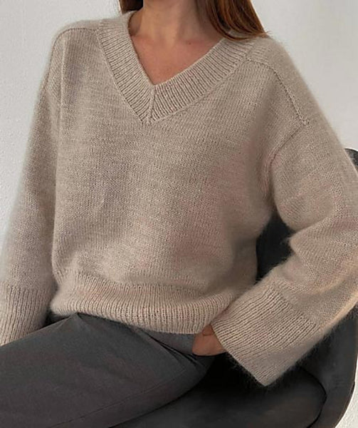 A person shown from the neck down, wearing a knit long sleeved sweater. They are sitting with one hand in their pocket. The other hand is not visible. A brown chair and white wall are visible behind the person.