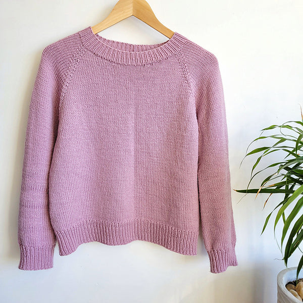 Pink handknit raglan pullover on a wooden hanger on a white background with a plant to the right