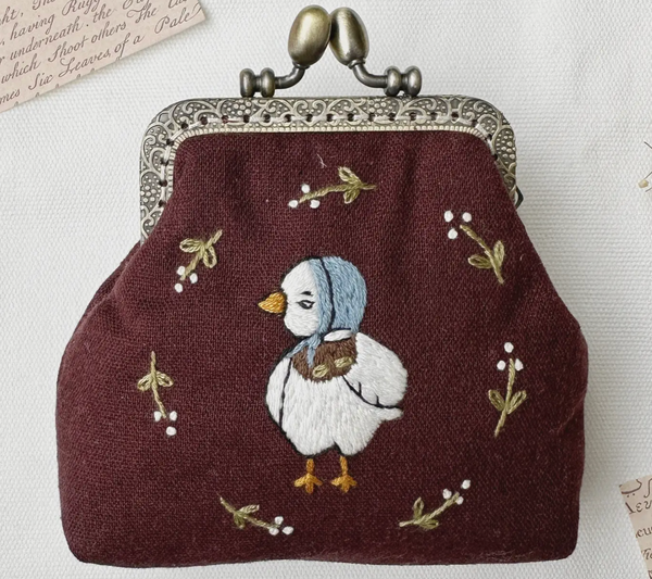 A small vintage-style kiss purse in deep burgundy fabric, featuring an embroidered white duckling wearing a blue bonnet, surrounded by delicate gold and white floral embroidery. The purse has an ornate silver-tone metal clasp at the top.