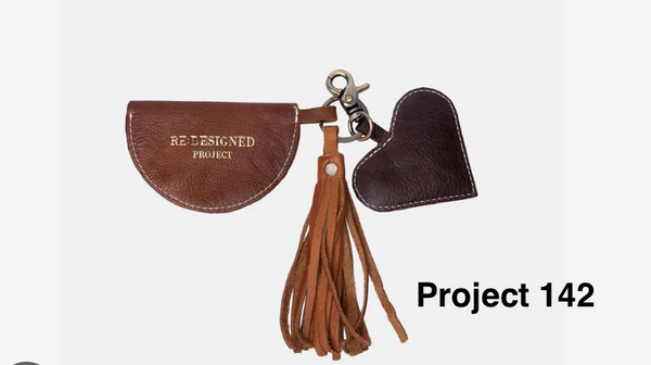 A brown leather keychain set featuring a semicircle pouch with gold embossed 'REDESIGNED PROJECT' text, a dark brown heart-shaped tag, and a tan tassel, shown on a light gray background with 'Project 142' text overlay.