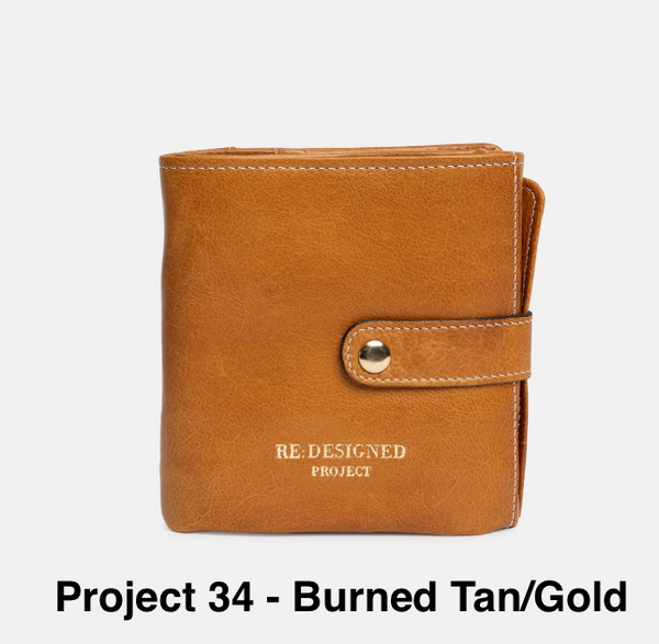 A tan leather wallet with a strap closure and gold-tone snap button, shown on a light gray background with the text 'Project 34 - Burned Tan/Gold' below it. The wallet has 'RE:DESIGNED PROJECT' printed on the front.