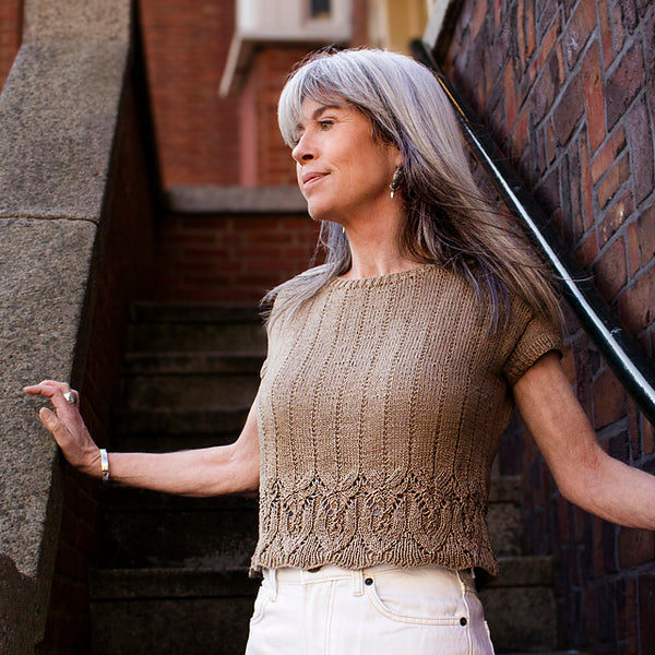 A woman standing on outdoor stairs wearing a short sleeved knitted top with lace border detail and simple rib. She is looking away from the camera with her hands touching the surrounding wall.