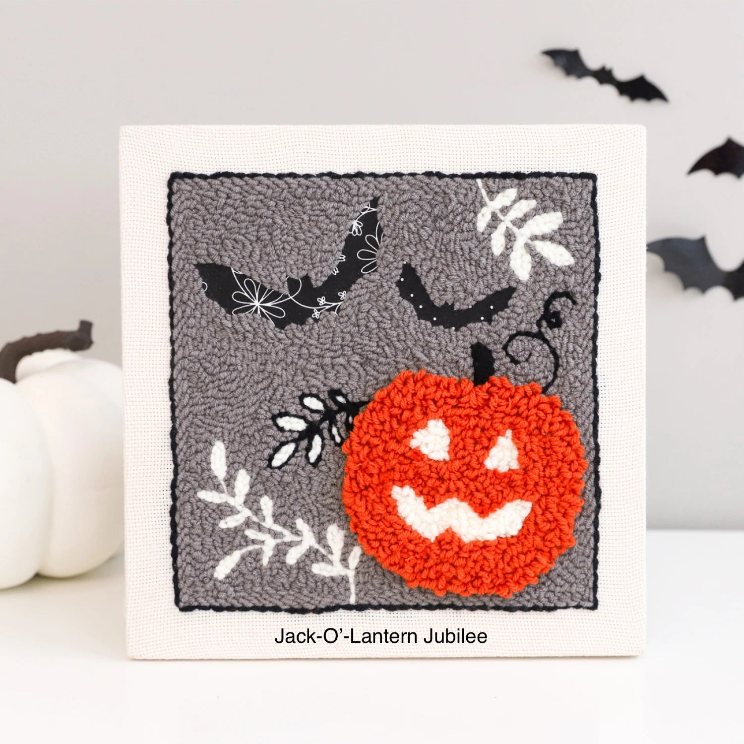 A 10"x10" cloth frame with a halloween theme punch needle design including pumpkin and fabric bats.
