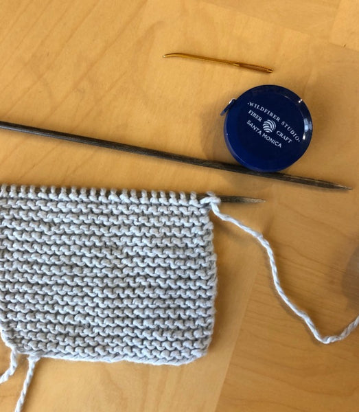 knitting needles with a tapestry needle and tape measurer. one knitting needle has a garter stitch piece of knitted work on it
