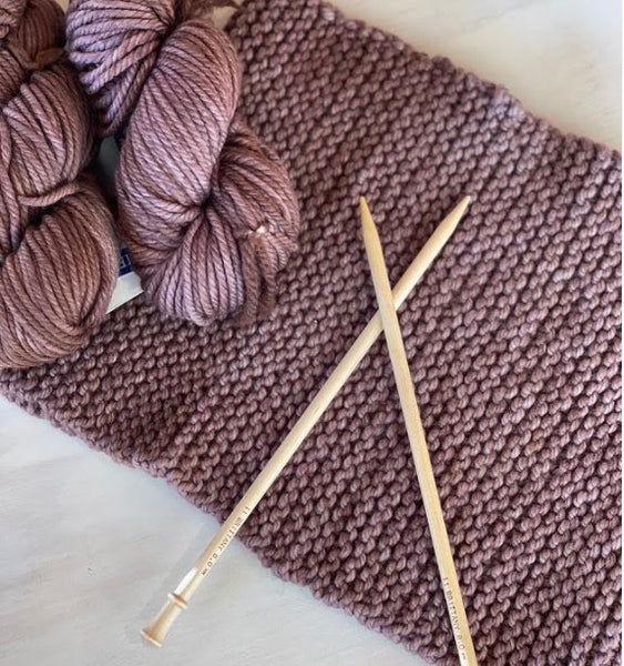 a knitted scarf with knitting needles on top and 2 skeins of yarn in light purple